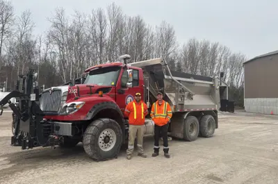 Two people in work wear stand in front of Township of Hamilton dump truck with chains on the wheels