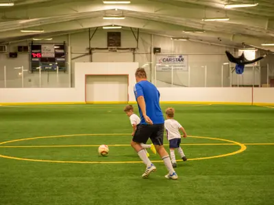 Person playing soccer with two small children at an indoor soccer arena