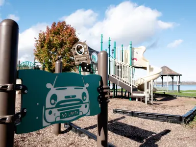 Optimist Park playground focused on a metal sign depicting a car with a license plate reading "no limits", behind the sign is a play structure and a gazebo close to Rice Lake