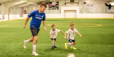 Person playing soccer with two small children at an indoor soccer arena