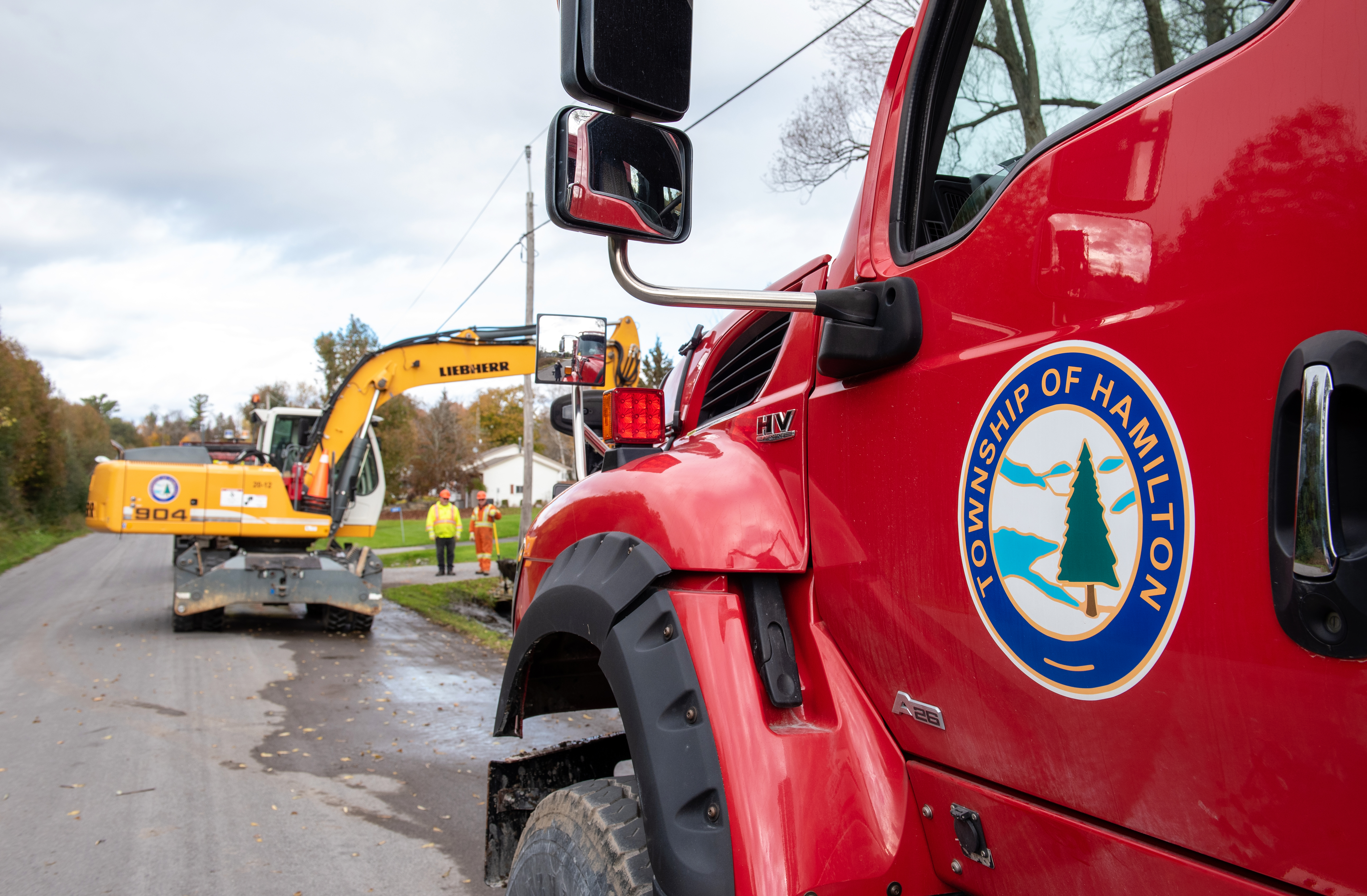 close up of a dump truck focused on the Township of Hamilton decal on the driver's side door, in the background there is an excavator and two people working on a ditch