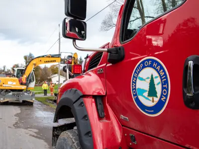 close up of a dump truck focused on the Township of Hamilton decal on the driver's side door, in the background there is an excavator and two people working on a ditch