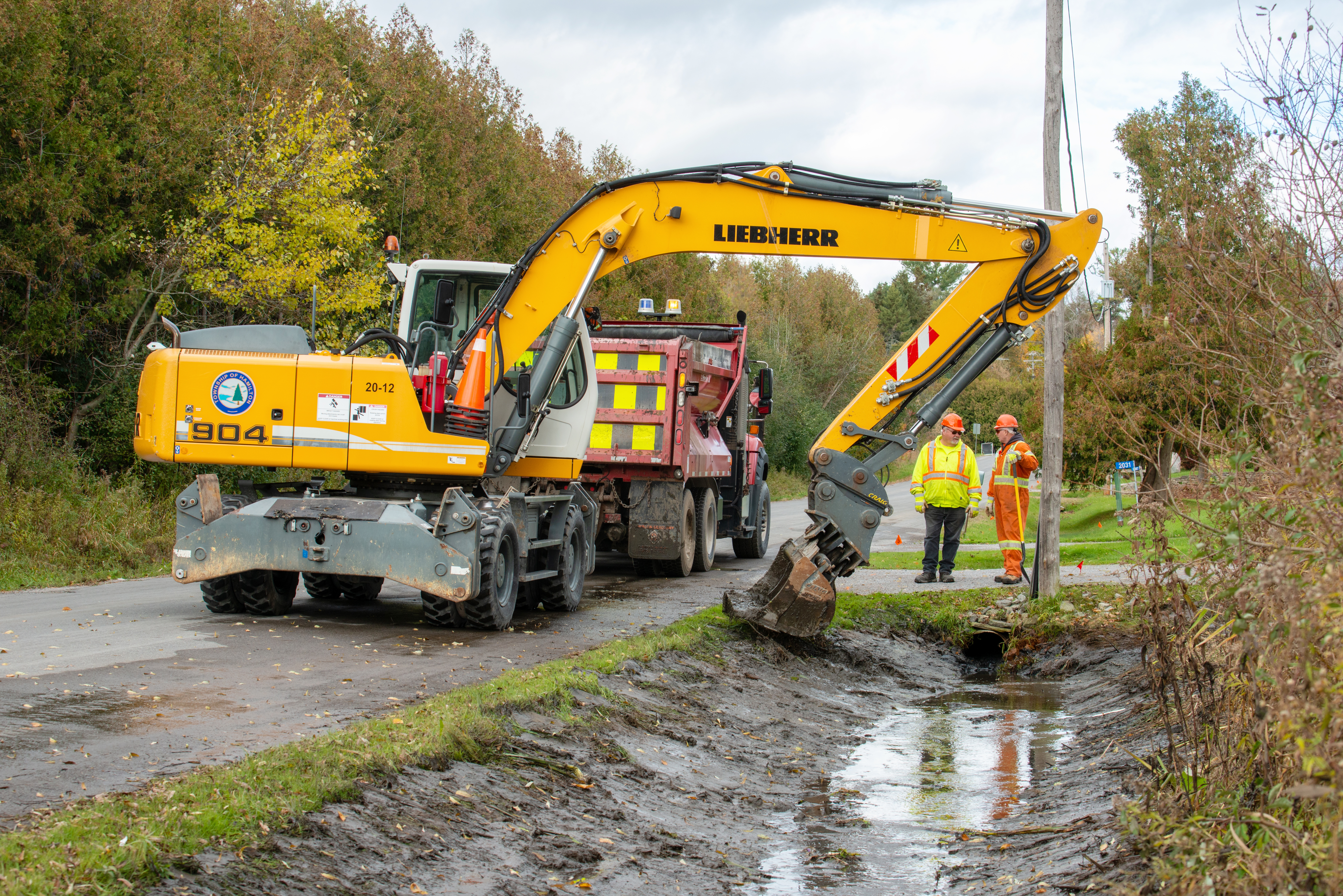 excavator digging in a ditch with two workers watching and a dump truck in the background
