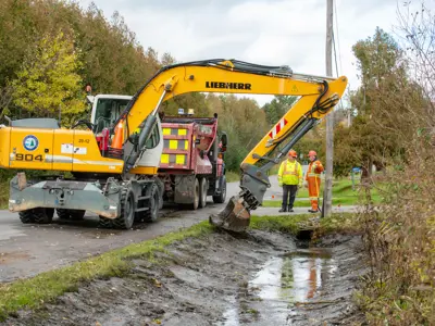 excavator digging in a ditch with two workers watching and a dump truck in the background