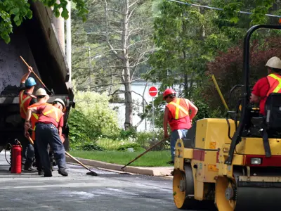 people working on a road, four are spreading asphalt from a dump truck whilst another is driving a road roller